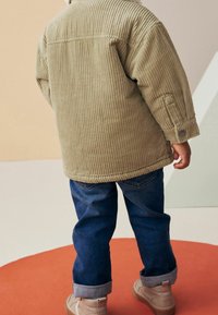 Veste en velours côtelé beige clair, avec une coupe décontractée et une texture côtelée, associée à un jean en denim bleu. Chaussures de teinte neutre visibles.