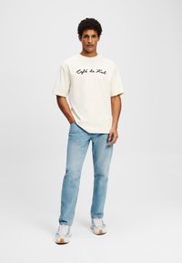 Cream oversized t-shirt with "Café de Zal" in black cursive, paired with light blue straight-leg jeans and white sneakers.