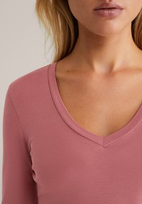 Hosszú ujjú, V-kivágású felső puha, poros rózsaszín pamutkeverékből. Sima textúra finom bordázással, minimalista dizájn, díszítések nélkül.
