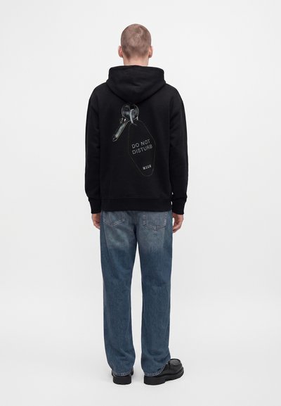 Personne portant un sweat à capuche noir avec un graphique de clé et une étiquette "DO NOT DISTURB" dans le dos, un jean bleu et des chaussures noires, debout de dos.