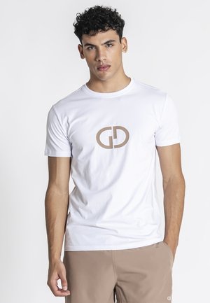 Mladić s kovrčavom crnom kosom, odjeven u bijelu majicu s bež logom "GD" i bež hlačama, stoji nasuprot jednostavnoj bijeloj pozadini.