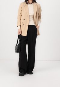 Femme portant un blazer beige, un pull côtelé crème, un pantalon large noir, des chaussures noires, tenant un sac à main rond noir clouté.