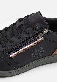 Adidași negri cu șireturi și fermoar pe lateral. Au accente în dungi maro și albe și o finisare texturată. Talpă din cauciuc gri.