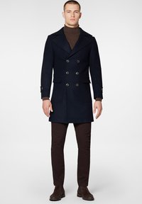 Navy wełniany płaszcz podwójnie zapinany na guziki z ostrymi klapami, dwoma przednimi kieszeniami i mankietami zapinanymi na guziki, zestawiony z brązowymi dopasowanymi spodniami i ciemnymi botkami.