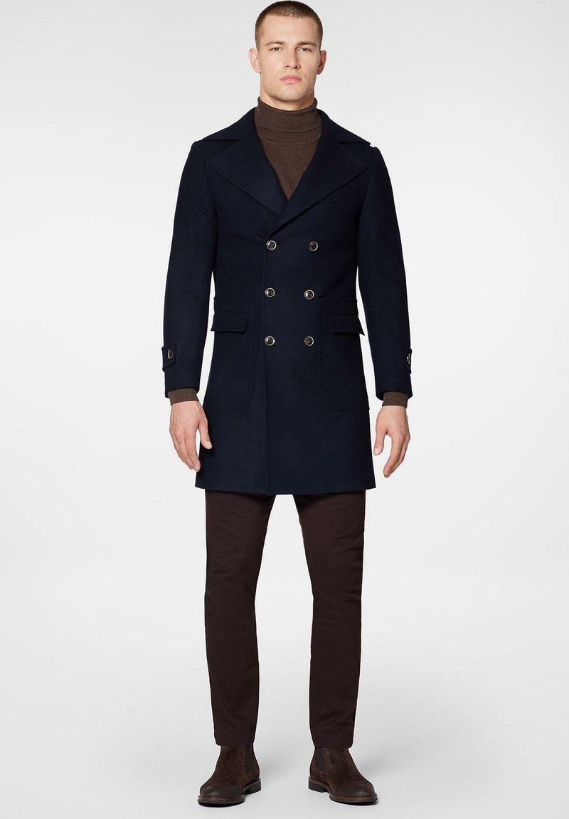 Navy wełniany płaszcz podwójnie zapinany na guziki z ostrymi klapami, dwoma przednimi kieszeniami i mankietami zapinanymi na guziki, zestawiony z brązowymi dopasowanymi spodniami i ciemnymi botkami.