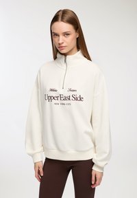 Biały sweter z wysokim kołnierzem, pół zamkiem i ściągaczami na mankietach. Zawiera bordowy haftowany napis "Upper East Side, Nowy Jork".