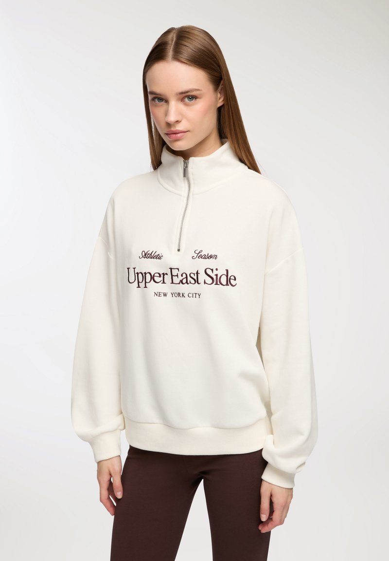 Biały sweter z wysokim kołnierzem, pół zamkiem i ściągaczami na mankietach. Zawiera bordowy haftowany napis "Upper East Side, Nowy Jork".