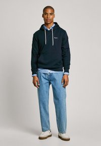 Sudadera navy con cordones, bolsillo delantero y logo blanco; camisa de botones azul claro debajo; jeans claros de corte relajado; zapatillas blancas.