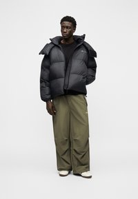 Νέος άνδρας που  φοράει μαύρο μπουφάν puffer, μαύρο πουκάμισο, άνετο χακί παντελόνι και λευκά αθλητικά παπούτσια, στέκεται μπροστά σε ένα απλό ανοιχτόχρωμο φόντο.