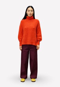 Vrouw met lang zwart haar, gekleed in een oranje coltrui, losse donkerpaarse broeken en gele schoenen, staande tegen een witte achtergrond.