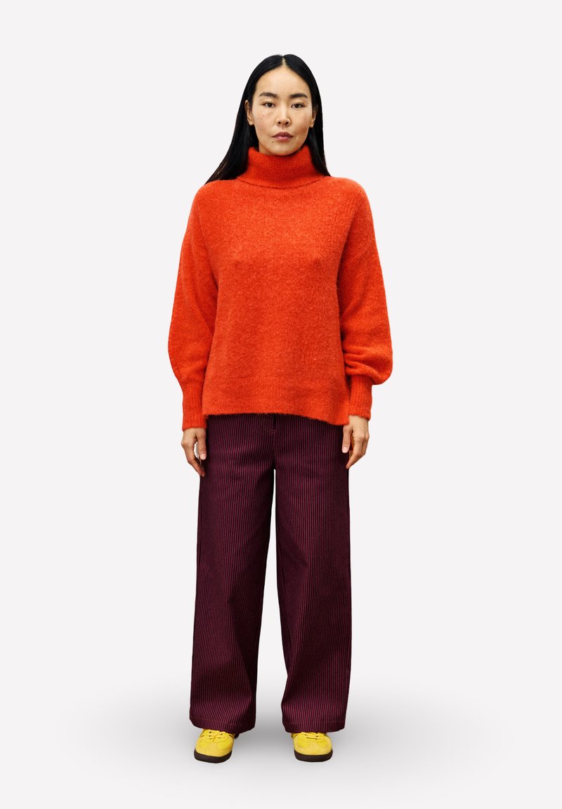 Vrouw met lang zwart haar, gekleed in een oranje coltrui, losse donkerpaarse broeken en gele schoenen, staande tegen een witte achtergrond.