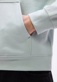 Χέρι που εισάγεται στην μπροστινή τσέπη ενός ανοιχτόγκρι φούτερ με ριμπ μανσέτα, δείχνοντας τον καρπό και μέρος από μαύρο παντελόνι από κάτω.