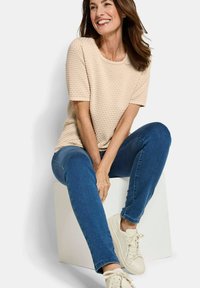 Een vrouw zit op een witte kubus, gekleed in een beige textuur met korte mouwen, blauwe jeans en witte sneakers. Ze glimlacht en kijkt naar rechts.