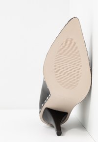 Musta nahkainen korkokengän malli, jossa on terävä kärki, hopeiset niitit reunassa ja teksturoitu beige pohja.