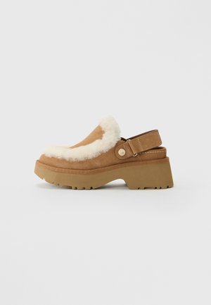 Zoccolo in suede beige con suola spessa, bordatura in shearling bianco e cinturino posteriore regolabile con dettaglio a bottone dorato su sfondo semplice.