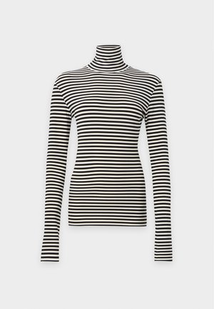 Jeune femme aux cheveux bouclés porte un col roulé beige et noir rayé et un pantalon évasé noir avec des rayures blanches verticales, debout sur fond blanc.
