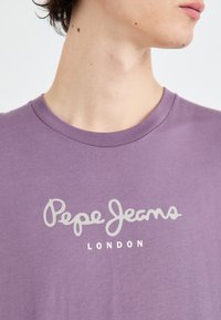 Violeta kokvilnas t-krekls ar apaļu kakla izgriezumu, īsām piedurknēm un baltiem drukātiem uzrakstiem "Pepe Jeans LONDON" krūtīs.