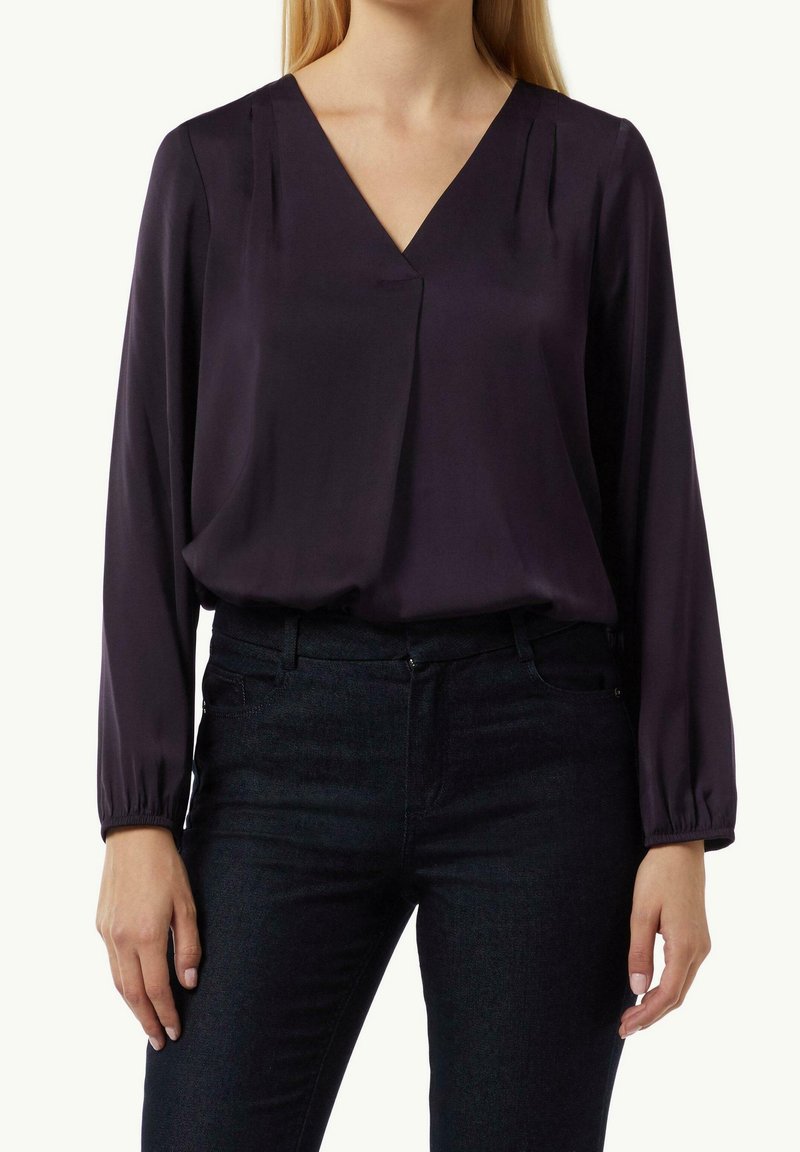 Blouse en satin violet avec col en V, manches longues, et plis subtils à l'avant, assortie à un jean foncé. Texture lisse et coupe décontractée.