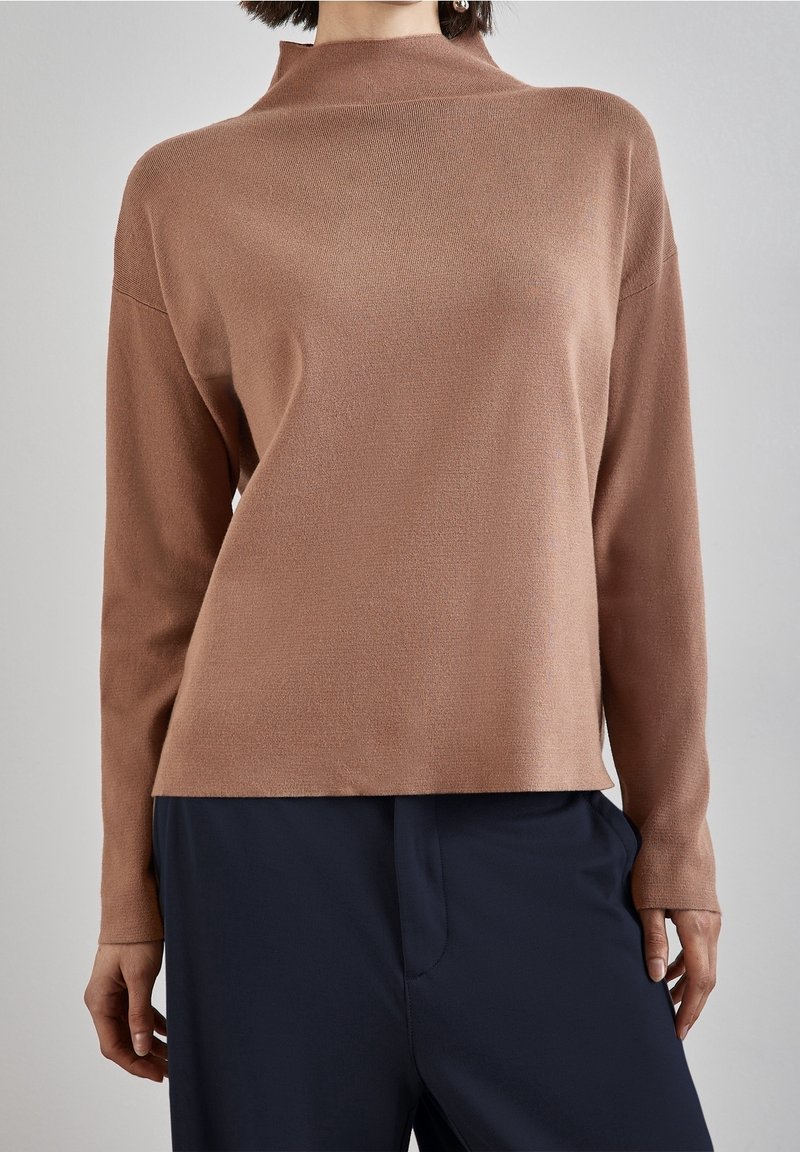 Brun stickad tröja med hög krage, långa ärmar och avslappnad passform. Slät textur och minimalistisk design. Matchad med mörka byxor.