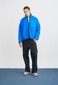 Niebieska polarowa kurtka z wysokim kołnierzem, zapinana na zamek, z kieszonką na piersi. W połączeniu z czarnymi spodniami i szarymi trampkami z żółtymi akcentami.