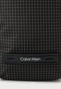 Fekete szövet hátizsák rácsmintával, sima bőrcímkével, amelyen a "Calvin Klein" felirat olvasható. Minimalista dizájn és textúra részletek.