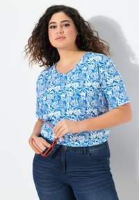 Modrý květinový top s krátkým rukávem a výstřihem ve tvaru V, zdobený bílými botanickými vzory; kombinovaný s tmavými džínami a červenými slunečními brýlemi.