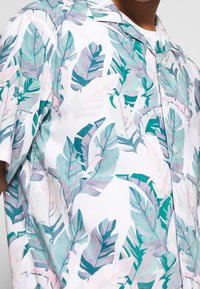 Kortärmad skjorta i bomull med vit bakgrund och färgglada palmbladstryck i turkosa, rosa och gröna nyanser. Knäppning med knappar.