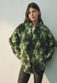 Pulover od flisa u zelenom kamuflažnom uzorku, s visokim ovratnikom i dizajnom s polu-zipom; meka tekstura i oversized silueta.