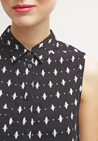 Czarna koszulka bez rękawów z białym geometrycznym wzorem, z szerokim kołnierzykiem i czarnymi guzikami z przodu. Gładka faktura materiału.