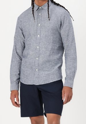 Homme portant une chemise boutonnée à manches longues gris clair et un short bleu foncé, debout, les bras détendus le long du corps.