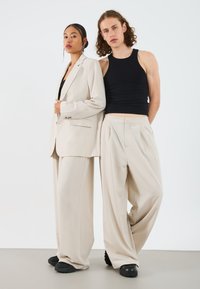 Une blazer oversized beige et un pantalon large sur une femme, associée à un débardeur noir ; un homme en débardeur noir et pantalon beige, tous deux en coupes décontractées.