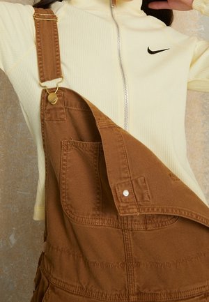 Persoon draagt een crèmekleurige Nike trui met rits onder een bruine denim tuinbroek met een bandje los, met gedetailleerd stikwerk en metalen sluitingen.