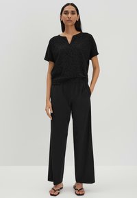 Zwarte short-sleeve top met een subtiel patroon en V-hals, gecombineerd met losse zwarte broek. Beide gemaakt van lichtgewicht stof. Platte sandalen.