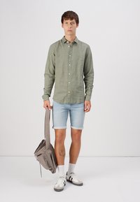 Camisa verde claro de botones, pantalones cortos de mezclilla, calcetines blancos, bolso gris de hombro. El modelo tiene el cabello corto y está de pie contra un fondo neutro.