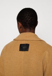 Personne aux cheveux courts bouclés portant un manteau couleur camel avec une étiquette noire « HUGO » cousue au col arrière.