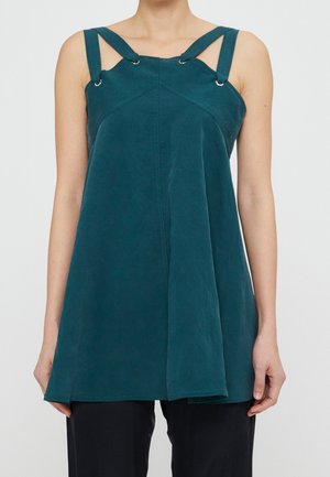Haut sans manches bleu sarcelle avec larges bretelles reliées par des anneaux métalliques, coupe ample, porté avec un pantalon noir, montré du cou aux hanches.