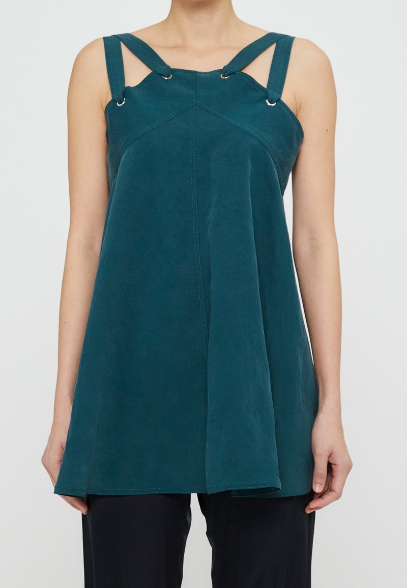 Haut sans manches bleu sarcelle avec larges bretelles reliées par des anneaux métalliques, coupe ample, porté avec un pantalon noir, montré du cou aux hanches.