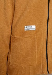 Senapsgul stickad tröja med dragkedja, utrustad med en sidoficka och en vit etikett med svart text. Mjuk textur och ribbade muddar.