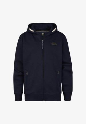 Marineblå hættetrøje med lynlås og hvidt foret hætte, med forlommer og et logo på brystet, kombineret med lyseblå jeans og hvide sko.