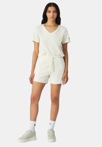 Creme V-hals T-shirt gecombineerd met bijpassende elastische taille shorts, beide gemaakt van zacht materiaal. Model draagt lichte sneakers en witte sokken met logo.