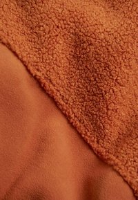 Rostfarbener Stoff, der zwei Texturen zeigt: einen glatten, matten Bereich und einen strukturierten, flauschigen Teil mit einer weichen, erhabenen Oberfläche.