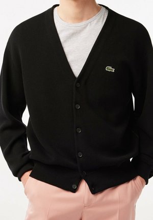 Personne portant un cardigan noir boutonné avec un logo crocodile vert sur une chemise gris clair et un pantalon rose clair, mains dans les poches.