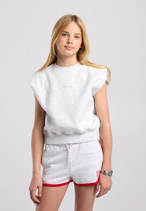 Tienermeisje met lang blond haar draagt een lichtgrijze mouwloze trui en grijze shorts met rode bies, staand tegen een effen achtergrond.