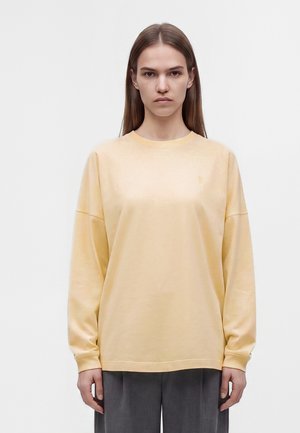 Donna con capelli castani lisci che indossa una camicia larga giallo pallido a maniche lunghe e pantaloni grigi su uno sfondo bianco uniforme.