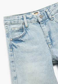 Vaqueros de denim azul claro con un aspecto desgastado, que cuentan con cierre de cremallera, bolsillos delanteros y costuras amarillas en las costuras.