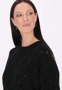 Schwarzer Strickpullover mit einem Zopfmuster, rundem Ausschnitt und subtilen Glitzerakzenten. Das Model trägt goldene Creolen.