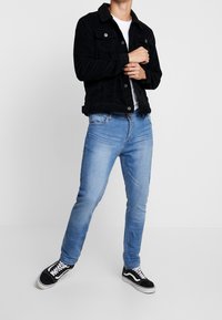 Man i svart jacka med sherpa-krage, vit t-shirt, ljusblå jeans och svarta sneakers, poserar mot en helt vit bakgrund.