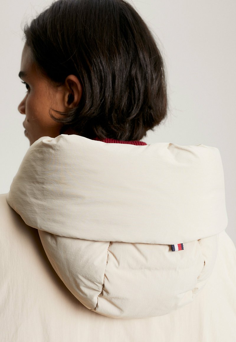 Beige gepolsterte Kapuze mit einem gesteppten Design, die an der Seite ein kleines dreifarbiges Etikett aufweist. Das Material wirkt weich und leicht glänzend.