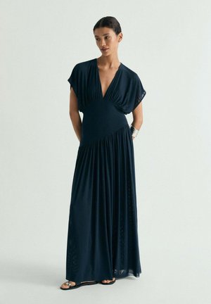 Mujer de pie con las manos detrás de la espalda, vistiendo un vestido maxi negro fluido con escote en pico profundo y brazalete de plata, sobre un fondo claro y liso.