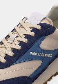 Κοντινή λήψη ενός αθλητικού παπουτσιού σε μπεζ, μπλε και ναυτικό μπλε χρώμα, με εκρού κορδόνια και το λογότυπο "KARL LAGERFELD" στο πλάι.